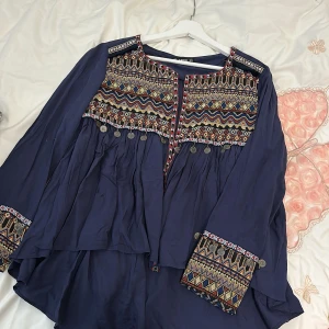 Mörkblå topp med broderade detaljer från Shein - Säljer en superfin mörkblå topp från Shein. Den har ett färgglatt broderat mönster över bröstet och ärmarna, med små hängande myntdetaljer som ger en bohemisk touch. Toppen är långärmad och har en lös passform, perfekt för en avslappnad stil.