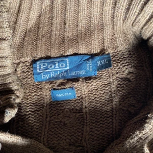 Polo Ralph Lauren stickad tröja - Säljer en snygg brun stickad tröja från Polo Ralph Lauren i storlek XXL. Tröjan är gjord av 100% silke och har ett klassiskt flätmönster. Den har långa ärmar och en hög krage med två knappar. Perfekt för höst och vinter! 