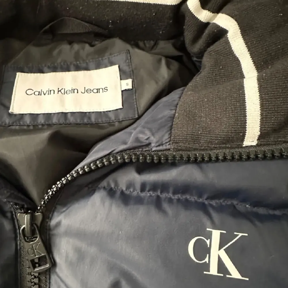 Säljer en snygg mörkblå pufferjacka från Calvin Klein Jeans. Jackan har en dragkedja framtill och en mysig krage med vita detaljer. Perfekt för kalla dagar med sin vadderade design. Den har också en avtagbar huva för extra skydd mot väder och vind.. Jackor.