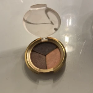 Ögonskugga i nyansen Brown Sugar - Säljer en elegant ögonskugga från Jane Iredale i nyansen Brown Sugar. Den kommer i en snygg guld- och transparent förpackning med tre olika färger: mörkbrun, ljusbrun och beige. Perfekt för en naturlig look eller för att skapa djupare ögonmakeup. Förpackning är lite sliten 