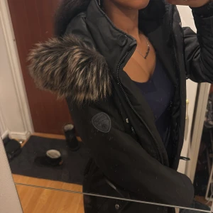 Svart parkajacka med pälsdetaljer - Säljer en snygg svart parkajacka med pälsdetaljer på huvan. Jackan har dragkedja och knappar framtill, perfekt för kalla dagar. Den är i mycket bra skick och passar perfekt för höst och vinter.❤️