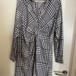 Svartvit mönstrad klänning från NA-KD - Snygg svartvit klänning från NA-KD i storlek 38. Den har ett coolt grafiskt mönster och långa ärmar. Perfekt för både vardag och fest! Klänningen har en knytning framtill som ger en fin passform. Nyskick , tvättad 1 gg. 