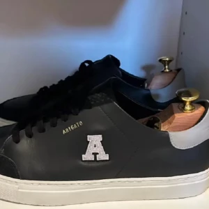 Svarta sneakers från Axel Arigato - Snygga svarta sneakers från Axel Arigato med en vit sula och ett stort 'A' på sidan. De har en stilren design med snörning och är perfekta för både vardag och fest. Skorna är i mycket bra skick och passar till alla outfits.
