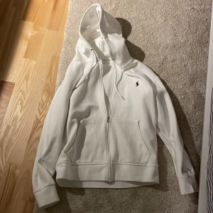 Vit hoodie från Ralph Lauren - Säljer en stilren vit hoodie från Ralph Lauren i storlek S. Den har en dragkedja framtill och den klassiska loggan broderad på bröstet. Perfekt för både vardag och chill dagar. Grymt skön och i mycket bra skick! 🏷️