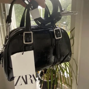 Svart handväska från Zara - Snygg svart handväska från Zara i nytt skick. Väskan är glansig med silverdetaljer och en justerbar axelrem. Köpt här på Plick! 🖤(första bilden lånad)