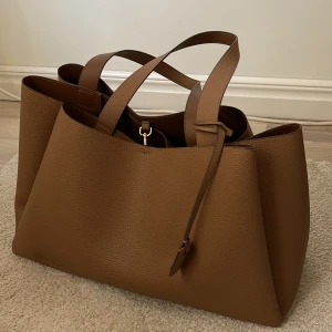 Brun axelväska - Säljer en stor snygg brun axelväska med stilren design. Väskan har två långa handtag och 3 praktiska innerfickor och fina guld detaljer. Helt ny och aldrig använd! Varan är i nytt skick men orginalförpackning följer inte med