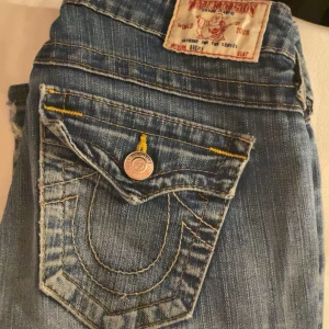 Blå jeans från True Religion w25 - Säljer ett par snygga blå jeans från True Religion i bra skick. De har en klassisk femficksdesign med coola sömmar och knappar. Jeansen är lågmidjade och har en bootcut-stil som ger en avslappnad look. Perfekta för både vardag och fest! Kan också tänka mig att byta mot ett par i storlek 24 eller 23❣️säljer pga förstora. Det är ett litet hål men syns inte så mycket och är lätt att sy igen