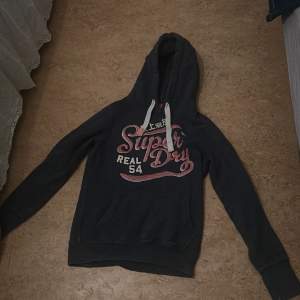 Säljer en svart hoodie från Superdry i bra skick. Den har en stor logga i rosa och vitt på framsidan och en mysig huva med vita snören. Perfekt för höst och vinter när du vill hålla dig varm och bekväm. 🖤 Storlek S men passar också Xs