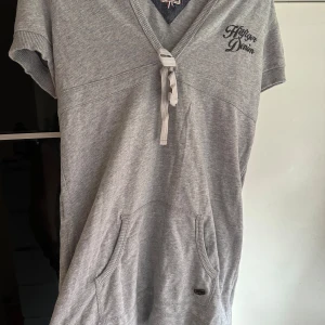 Grå hoodie från Hilfiger Denim - Säljer en snygg grå hoodie från Hilfiger Denim. Den har korta ärmar och en praktisk ficka framtill. Perfekt för en avslappnad stil. Den har en knytning vid halsen och en liten logga på bröstet. Superbekväm och stilren!