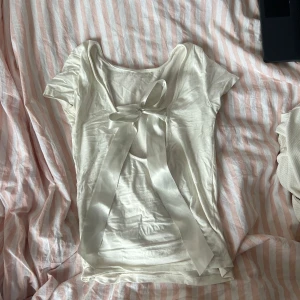 Vit kortärmad topp med rosett - Säljer en söt vit topp med korta ärmar och en stor rosett i ryggen. Toppen är i ett mjukt och bekvämt material som känns skönt mot huden.