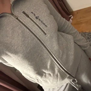Grå hoodie från Sail Racing - Säljer en snygg och bekväm grå hoodie från Sail Racing i storlek M. Den har en dragkedja framtill och en bröstficka med lite utrymme. Använd ett par gånger men i ett bra skick ☺️