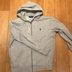 Grå hoodie från Polo Ralph Lauren - Säljer en stilren grå hoodie från Polo Ralph Lauren med dragkedja och det klassiska logotypbroderiet på bröstet. Tröjan har en justerbar huva och långa ärmar, perfekt för en avslappnad look.