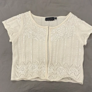 Vit kortärmad kofta från Vintage Stories - Charmig vit kofta från Vintage Stories med korta ärmar och vackra paljettbroderier. Perfekt för att ge en elegant touch till din outfit. Koftan är öppen framtill och har ett delikat mönster som ger en vintagekänsla.