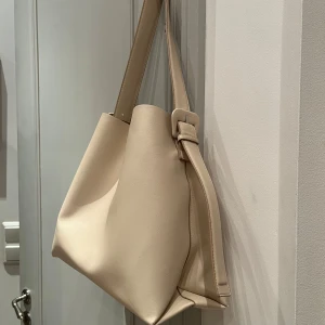 Beige axelväska med justerbart spänne - Snygg beige axelväska i syntetmaterial med ett stilrent och minimalistiskt utseende. Väskan har ett justerbart spänne på axelremmen och är köpt på mango förra säsongen.