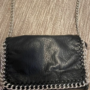 Svart axelväska med kedjedetaljer - Snygg svart axelväska i skinn med silverfärgade kedjedetaljer runt kanterna. Väskan har en dragkedja och magnetknappar för säker förslutning. Perfekt för att ge en edgy touch till din outfit.
