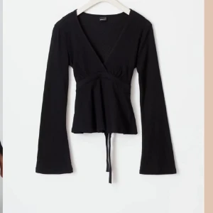 Svart ribbad topp  - Snygg svart ribbad topp från Gina med v-ringning och långa ärmar. Toppen har en knytning i midjan som ger en fin passform. 