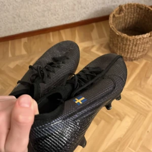 Nike mercurial vapor 13 - Säljer ett par svarta fotbollsskor med snörning och svensk flagga på sidan. Skorna har en texturerad yta och dobbar för bra grepp på planen. Perfekta för fotbollsspelare som vill ha stil och funktionalitet. (Flaggan går att ta bort)
