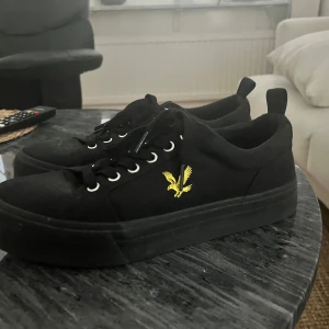 Svarta sneakers med gul detalj - Snygga svarta sneakers med en gul broderad fågel på sidan. Skorna har snörning och en tjock sula för extra komfort. Perfekta för en stilren look.