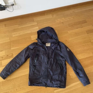 Mörkblå vindjacka från Moncler - Snygg mörkblå vindjacka från Moncler med huva och dragkedja. Jackan har Moncler-logga på ärmen och märkesnamnet tryckt längs dragkedjan. Perfekt för blåsiga dagar.