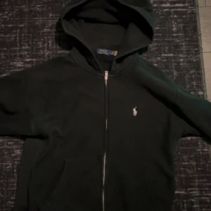 Svart huvtröja från Polo Ralph Lauren - Säljer en svart huvtröja från Polo Ralph Lauren med dragkedja och broderad logga på bröstet. Perfekt för en casual look med sin klassiska design och bekväma passform.