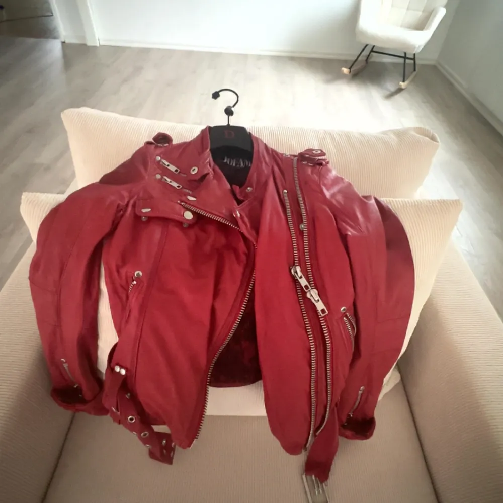 Snygg röd skinnjacka från Jofama med coola dragkedjedetaljer och knappar. Jackan har en tight passform och är perfekt för att ge din outfit en edgy touch. Den har långa ärmar och ett bälte i midjan för extra stil. Jackan har märken i bak, vet ej vad det är ifrån, därav priset. . Takit.