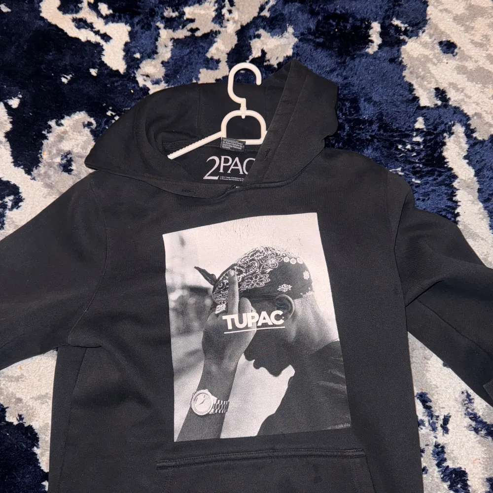 Snygg svart hoodie med ett stort tryck av Tupac på framsidan. Perfekt för dig som vill ha en bekväm och stilren tröja med en ikonisk design. Hoodien har en klassisk passform och en praktisk ficka framtill.. Neuletakit & Villapaidat.