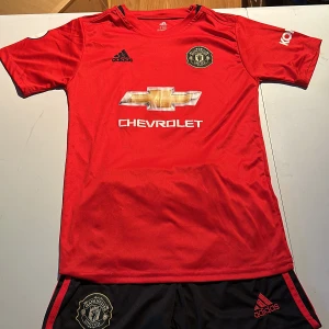 Röd Manchester United fotbollströja och shorts  - Säljer en röd Manchester United fotbollströja med shorts med Chevrolet-logga på framsidan. Tröjan har korta ärmar och klubbmärket på bröstet. Namnet 'Rashford' och nummer 10 är tryckt på baksidan. Perfekt för fans av laget!