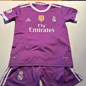 Lila Real Madrid fotbollströja och shorts - Säljer en lila Real Madrid fotbollströja med korta ärmar och matchande shorts. Tröjan har vit text och detaljer, inklusive 'Fly Emirates' och klubbens emblem. Baksidan visar 'Ronaldo 7'. Storlek ca 140. Perfekt för fans! 💜⚽