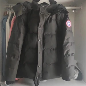 Svart dunjacka från Canada Goose - Säljer en svart dunjacka från Canada Goose med ikonisk logga på ärmen. Jackan har en praktisk dragkedja och knappar framtill, perfekt för kalla vinterdagar. Tillverkad i Kanada med högkvalitativa material.