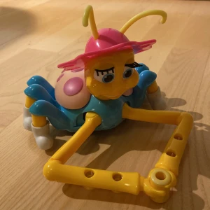 Leksak - Färgglad leksak i form av en insekt med rosa, blå och gula detaljer. Den har en hatt med stjärnor och cirklar som kan användas som en rit-mall. Leksaken har stora ögon och antenner, samt skor med olika mönster. Ska kunna rita av sig själv om man sätter dit pennor men den funktionen fungerar tyvärr inte längre därav lågt pris.