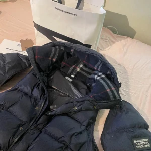 Svart dunjacka från Burberry - Snygg svart dunjacka från Burberry med klassiskt rutigt foder i huvan. Jackan har dragkedja och knappar framtill för extra skydd mot kyla. Perfekt för kalla dagar.
