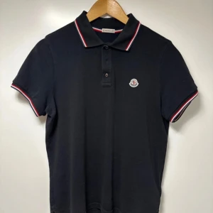 Svart Moncler Pikétröja - Snygg svart pikétröja från Moncler med korta ärmar. Den är nästan helt ny med inga märken på användning. Size M