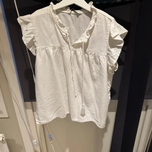 Vit blus med volangdetaljer - Charmig vit blus med volangdetaljer vid halsen och korta ärmar. Blusen har en knytning framtill och är tillverkad i ett lätt och luftigt material. Perfekt för en avslappnad och stilren look.