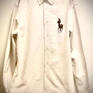 Vit långärmad Ralph Lauren skjorta med broderad detalj i storlek XL - Snygg vit långärmad skjorta med en broderad polospelare på bröstet. Skjortan har klassisk krage och knappar framtill. Perfekt för en stilren look. 