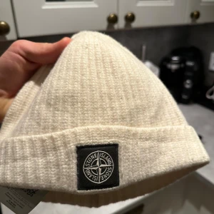 Vit stickad mössa från Stone Island - Snygg vit stickad mössa från Stone Island med ribbad design och det ikoniska märket framtill. Perfekt för att hålla värmen under kyliga dagar.