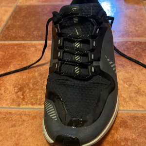 Svarta Nike sneakers storlek 38,5 - Snygga svarta Nike sneakers med snörning och vita detaljer. Perfekta för träning eller vardagsbruk. Skorna har en slitstark sula och en bekväm passform. Storlek 38,5. Ingen paket