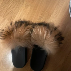 Svarta tofflor med pälsdetalj - Snygga svarta tofflor med fluffig pälsdetalj i beige och brun. Perfekta för att hålla fötterna varma och stiliga hemma.