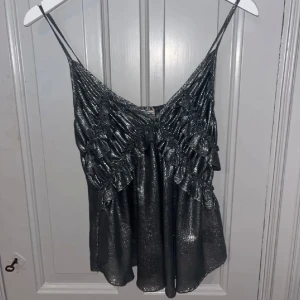 Silver topp med volanger - Lägger upp igen, snygg silver topp med smala axelband och volangdetaljer. Perfekt för en ute kväll med sitt skimrande material. Passar mig som är en xs/s normalt. Köpt vintage så väldigt ovanlig inte sett någon ha samma topp!