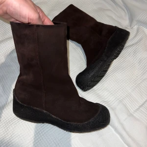 Bruna höga Bally stövlar - Snygga bruna stövlar i mocka med en praktisk dragkedja på sidan. De har en robust svart sula som ger bra grepp. Fodrade med fårull så dom håller värmen oavsett väder. Perfekta för höst- och vinterdagar.