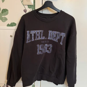 Svart sweatshirt från Gina Tricot - Säljer en svart sweatshirt från Gina Tricot med tryck 'ATHL. DEPT 1983' i blått och vitt. Tröjan har långa ärmar och en rund halsringning. Perfekt för en avslappnad stil.