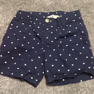 Mörkblå shorts med hjärtmönster - Säljer ett par mörkblå shorts med sött vitt hjärtmönster. De har en knapp och dragkedja framtill samt fickor både fram och bak. Perfekta för en avslappnad sommarlook!