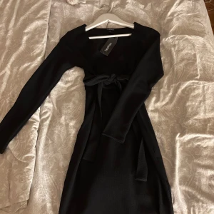 Svart klänning från Boohoo - Elegant svart klänning från Boohoo med långärmad design och knytband i midjan. Klänningen har en v-ringad omlottstil som ger en stilren look. Perfekt för både vardag och festliga tillfällen. Jätte fin klänning från Boohoo! Aldrig använd prislappen sitter kvar!  Nypris 350:-  Mitt pris 300:- eller bud