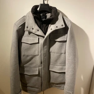 Grå Kappa från Jack and Jones - Snygg grå kappa från Jack and Jones. Jackan har en hög krage och stängs med dragkedja och knappar. Perfekt för lite kallare dagar! ✨Skick 8/10✨
