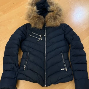 Svart dunjacka med päls från Hollies - Snygg svart dunjacka från Hollies med avtagbar päls på huvan och leopardmönstrat foder. Jackan har flera dragkedjefickor och en figursydd passform. Perfekt för kyliga dagar.