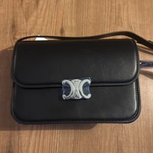 Svart axelväska med metallspänne - Snygg svart axelväska i skinn med ett elegant metallspänne framtill. Perfekt för att ge en stilren touch till din outfit. Väskan har en justerbar axelrem och är idealisk för både vardag och speciella tillfällen.