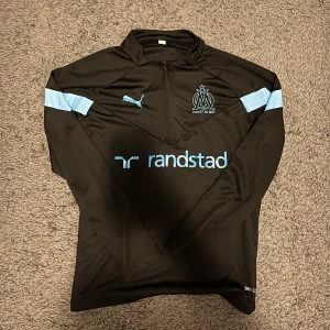 Svart träningsset från Puma - Säljer ett svart träningsset från Puma med Olympique de Marseille-logga. Tröjan har lång ärm och byxorna har elastisk midja. Båda delarna har ljusblå detaljer och Puma-logga. Perfekt för fotbollsträning eller vardagsbruk.