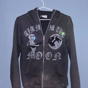 Svart hoodie med paljetter från Aim for the Moon - Snygg svart hoodie med dragkedja och paljettdekorationer från Aim for the Moon. Designen har en cool text och en figur på framsidan. Perfekt för en avslappnad stil med en touch av glitter.