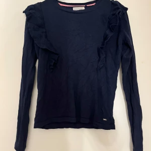 Mörkblå  tröja med volangdetaljer - Snygg mörkblå tröja med volangdetaljer längs axlarna. Tröjan har långa ärmar och en rund halsringning, perfekt för en stilren look.