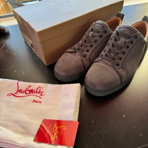 Gråa Christian Louboutin skor - Snygga grå sneakers från Christian Louboutin med klassisk röd sula. Skorna har snörning och är tillverkade i mocka, vilket ger en lyxig känsla. Perfekta för den som vill ha stilrena och exklusiva skor.