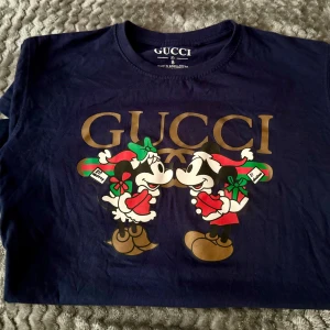 Gucci Mickey & Minnie Mouse ’21 Christmas t-shirt - Snygg blå t-shirt från Gucci med ett festligt julmotiv av musse & mimmi pigg i tomtekläder. Perfekt för att sprida julstämning! Stor logga i guld ovanför motivet. Har du någonsin känt behovet av att flexa på tomten? Han dyker upp varje år med sin släde, i år dyker du upp i Gucci. Gucci>Släde, det är sedan gammalt. Tomtemor kommer vara i dina DMs innan annandagen.